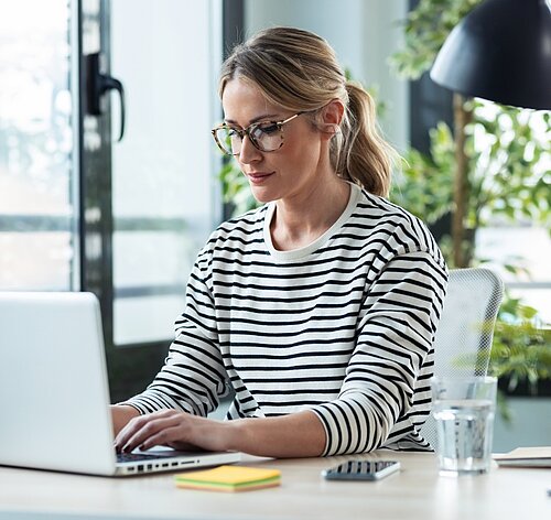 Eine Frau mit Brille tippt konzentriert an einem Laptop in einem hellen Arbeitsraum.  Das Bild zeigt eine Frau, die an einem Schreibtisch sitzt und an einem Laptop arbeitet. Sie trägt eine schwarz-weiß gestreifte Bluse und eine Brille mit gemustertem Rahmen. Ihre blonden Haare sind in einem lockeren Pferdeschwanz zusammengebunden. Ihr Blick ist auf den Laptop gerichtet, und ihre Hände tippen auf der Tastatur. Auf dem Schreibtisch befinden sich neben dem Laptop auch ein Stapel gelber Haftnotizen, ein Smartphone und ein Glas Wasser. Im Hintergrund sind ein Fenster mit schwarzen Rahmen, ein Schreibtischlampe und grünes Blattwerk einer Pflanze zu erkennen. Die Atmosphäre wirkt ruhig und konzentriert, das Bild vermittelt den Eindruck eines produktiven Arbeitsplatzes. Das Licht ist hell und natürlich, was die Szene freundlich und einladend wirken lässt.
