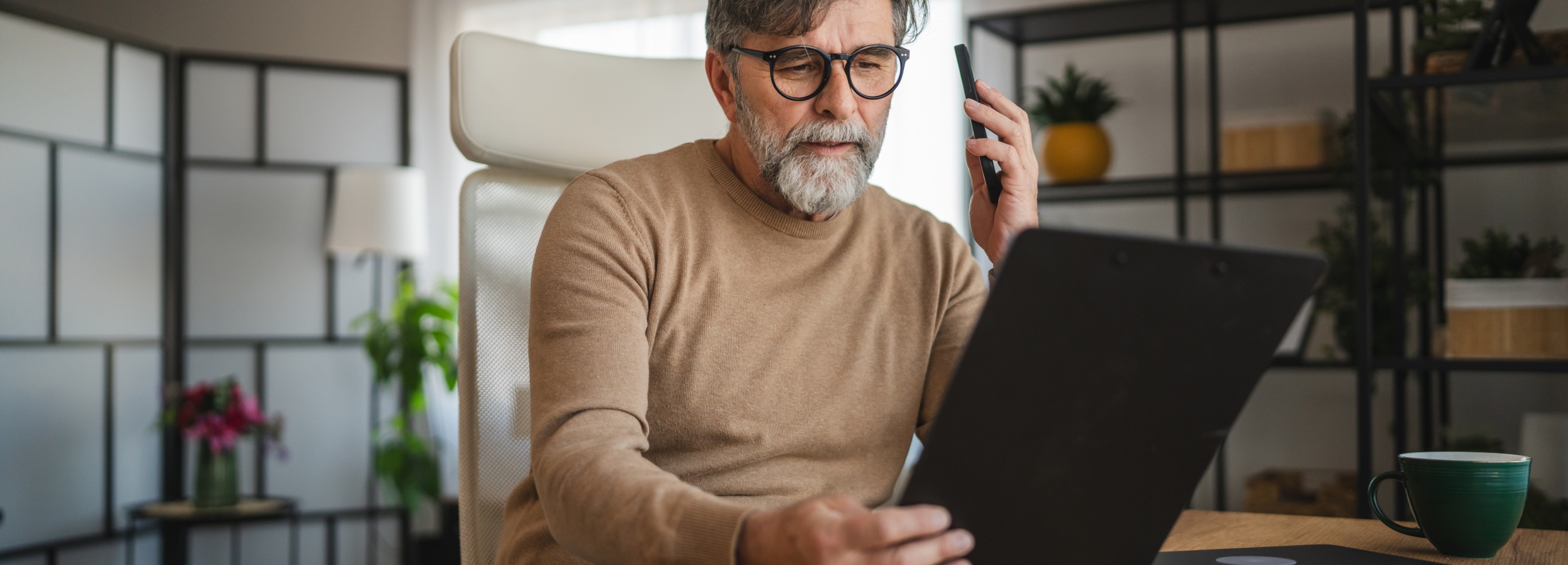 Ein älterer Mann sitzt mit Smartphone und Tablet an einem Schreibtisch und arbeitet.
